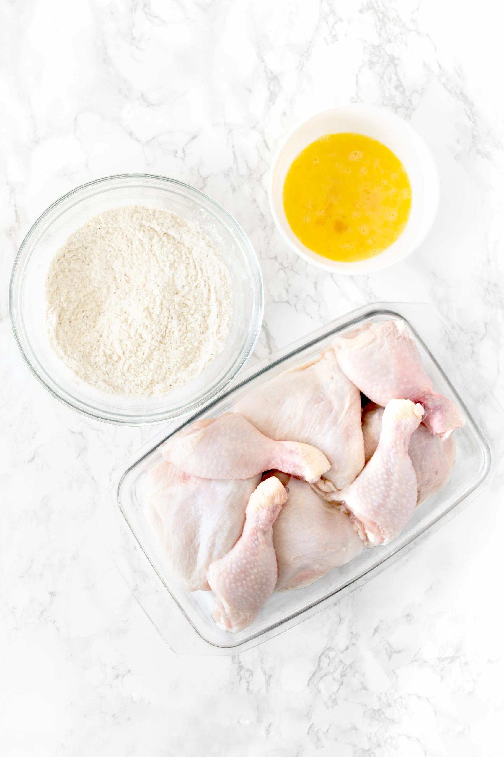 chicken, flour mixture, and beaten eggs for fried chicken on a white marble counter