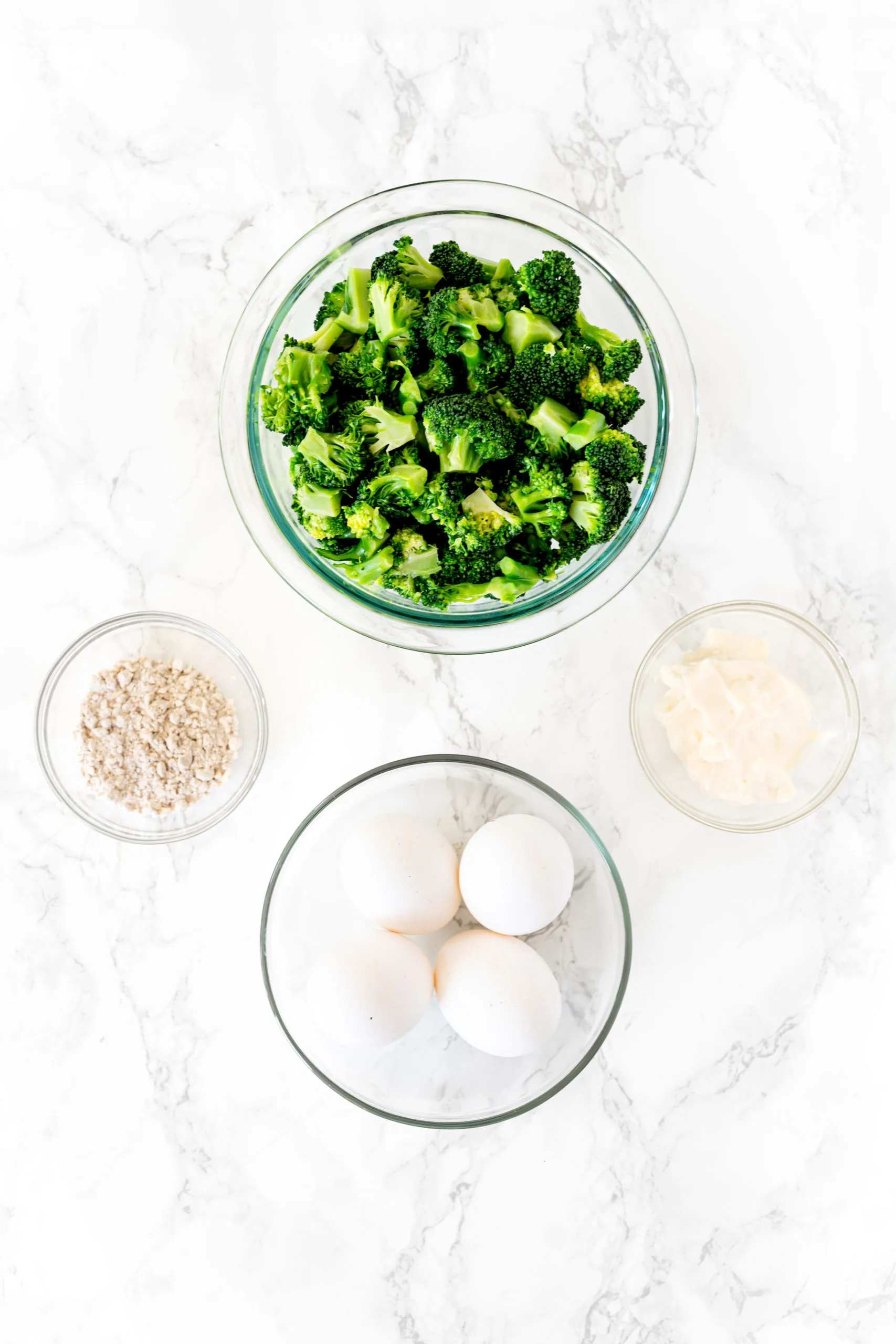 ingredients for broccoli kugel in bowls on a white marble counter