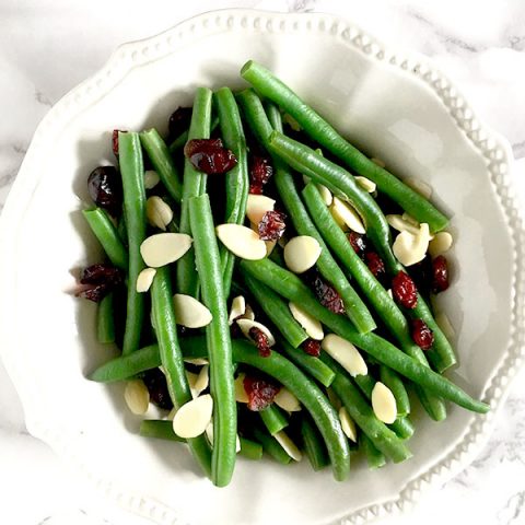 Green beans, cranberries, and dried almonds in a white bowl sitting on a white marble counter