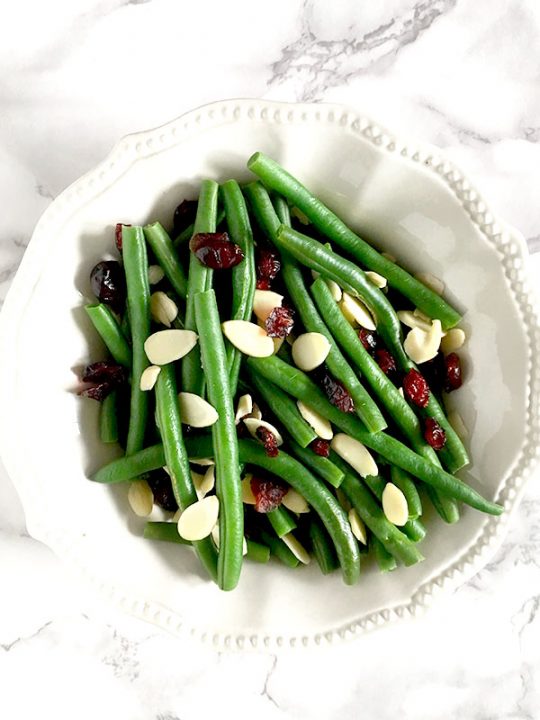 Green beans, cranberries, and dried almonds in a white bowl sitting on a white marble counter