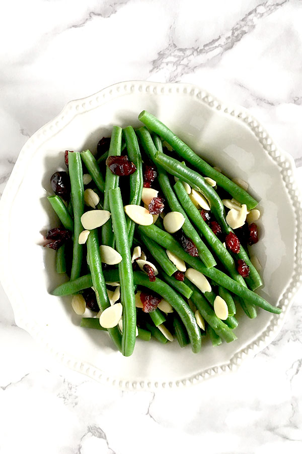 Green beans, cranberries, and dried almonds in a white bowl sitting on a white marble counter