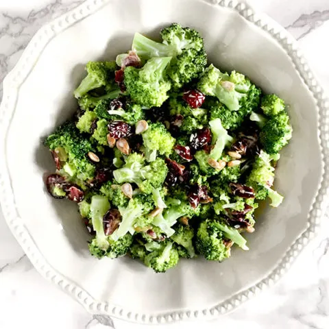 broccoli salad with cranberries and sunflower seeds in a white bowl on a white marble counter