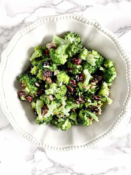 broccoli salad with cranberries and sunflower seeds in a white bowl on a white marble counter