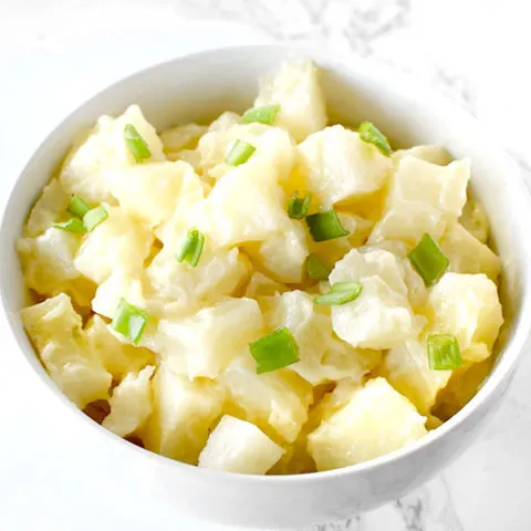 A simple potato salad topped with chives in a white bowl on a white marble counter