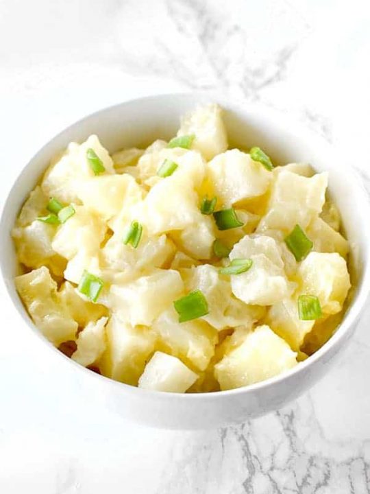 A simple potato salad topped with chives in a white bowl on a white marble counter