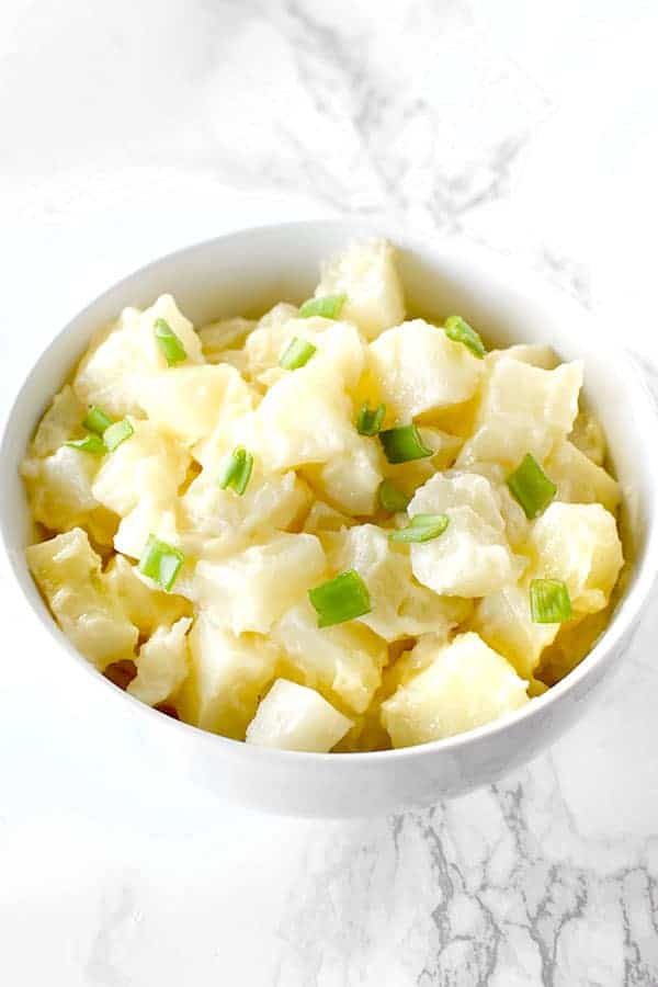 A simple potato salad topped with chives in a white bowl on a white marble counter