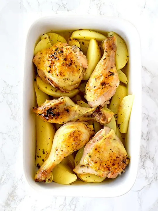 Greek lemon chicken and potatoes in a ceramic pan on a white marble counter