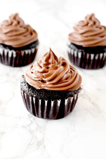 Three chocolate cupcakes with chocolate frosting on a white marble counter