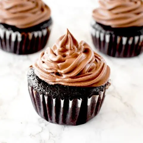 Three chocolate cupcakes with chocolate frosting on a white marble counter
