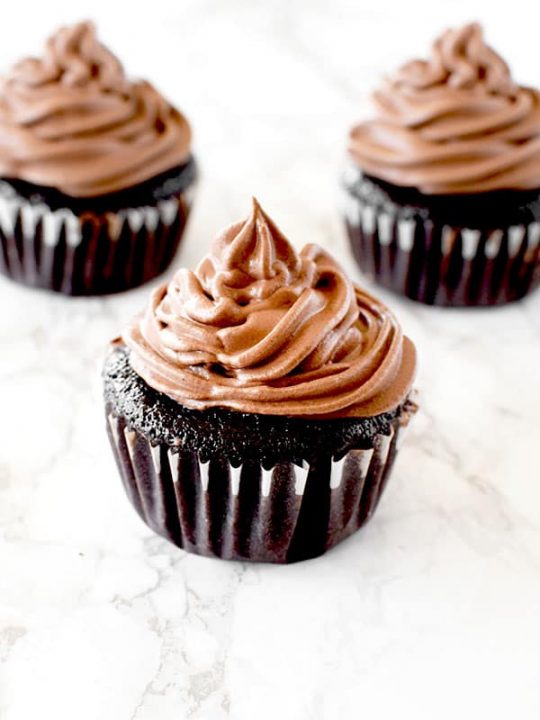 Three chocolate cupcakes with chocolate frosting on a white marble counter