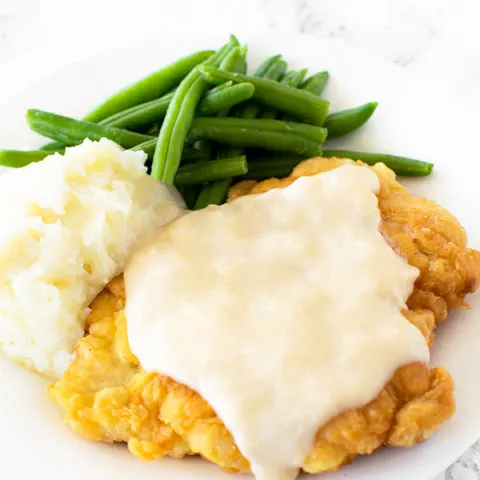 Chicken fried chicken with mashed potatoes and green beans on a white plate on a white marble counter