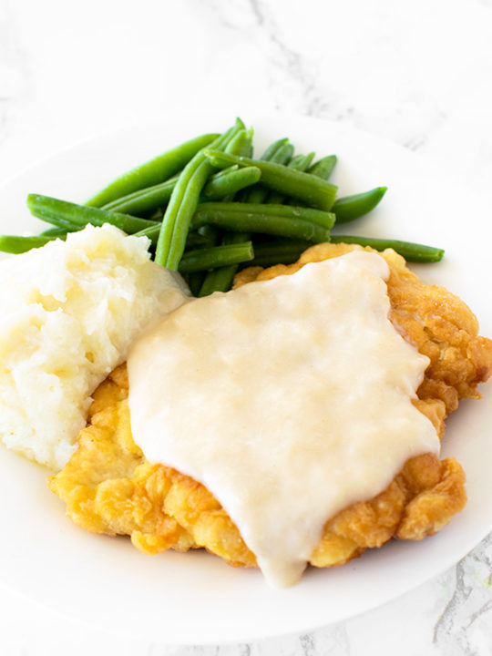 Chicken fried chicken with mashed potatoes and green beans on a white plate on a white marble counter