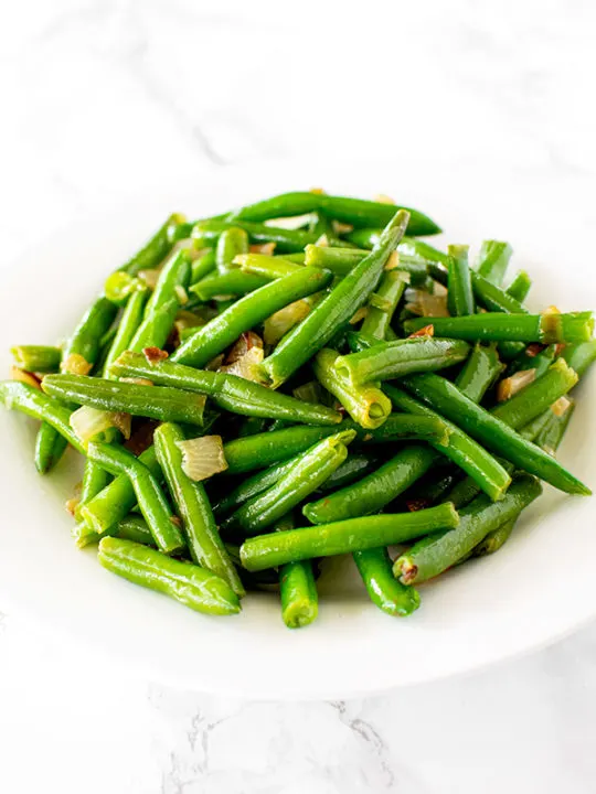 sauteed green beans with onions on a white plate on a white marble counter