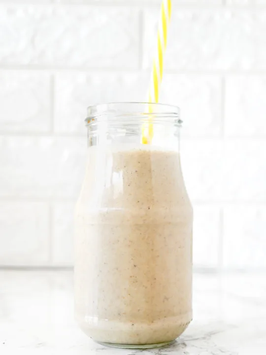 Banana Smoothie with Oat Milk in a glass with a yellow straw on a white marble counter