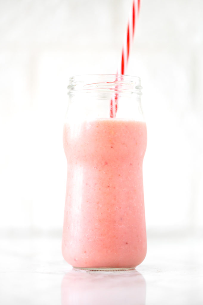Strawberry banana smoothie with coconut milk in a glass with a red straw on a white marble counter
