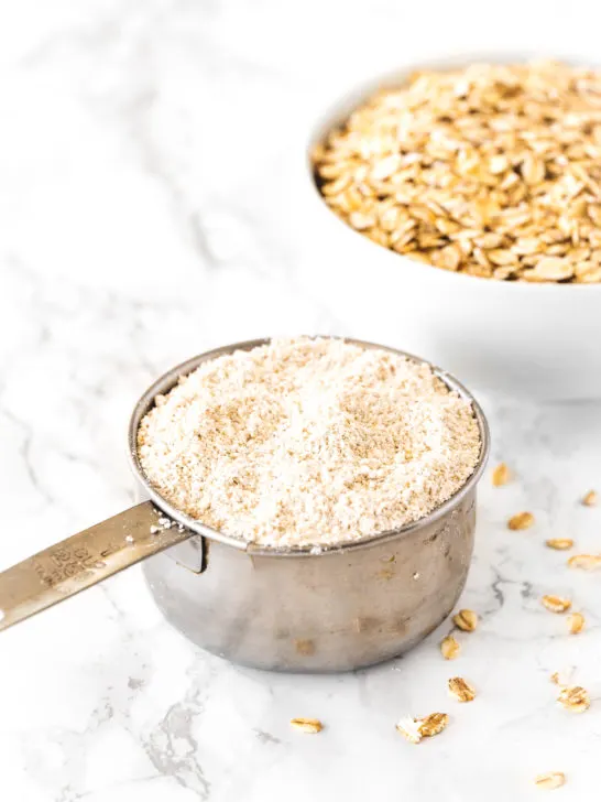 Oat flour in a measuring cup with a bowl of oats on a white marble counter