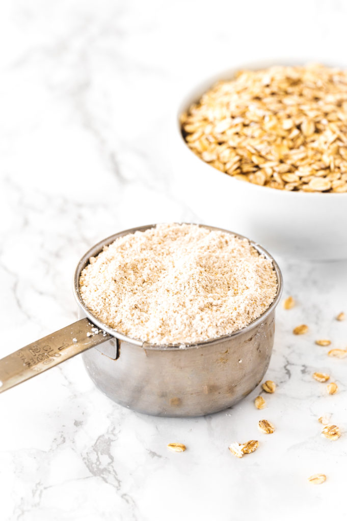 Oat flour in a measuring cup with a bowl of oats on a white marble counter