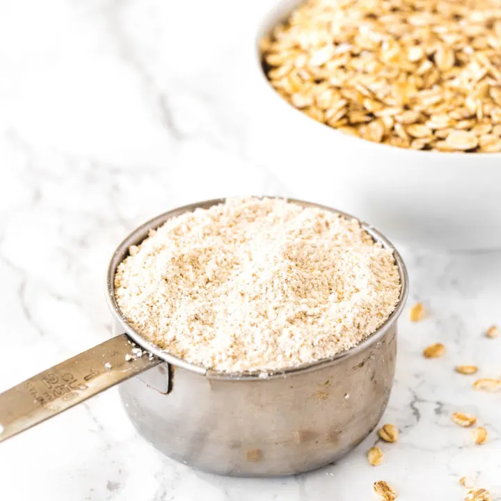 Oat flour in a measuring cup with a bowl of oats on a white marble counter